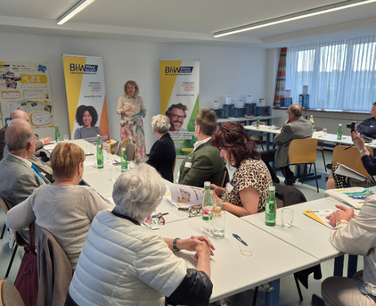 Foto von hinten von 3 langen Tischen welche in einer U Form aufgestellt sind. Alle Teilnehmer der Hauptregionskonferenz sitzen an diesen Tischen und schauen nach vorne zu der Vortragenden Person. Ebenso befinden sich Zettel und Getränke auf den Tischen.