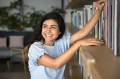 Eine lächelnde Frau greift in einem hellen Raum nach einem Buch aus einem vollen Bücherregal. Im Hintergrund sind Pflanzen und Sitzmöbel zu sehen, die eine gemütliche, inspirierende Atmosphäre schaffen.