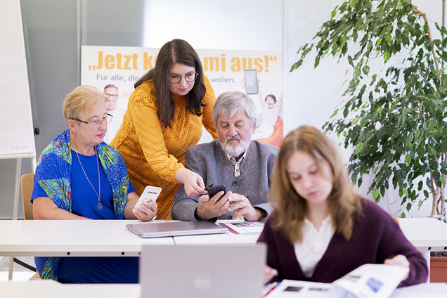 Eine junge Frau unterstützt zwei ältere Personen beim Umgang mit dem Smartphone, während eine Schülerin im Vordergrund konzentriert in den Lehrunterlagen blättert. Die Szene spielt sich in einem modernen Lernumfeld ab, das digitale Bildung für alle Generationen fördert.