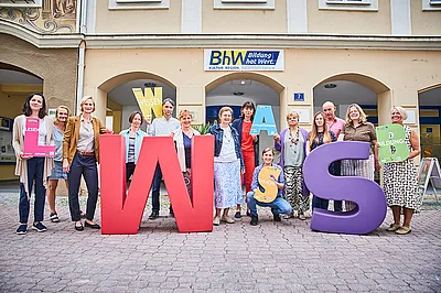 Die Lernbegleiterinnen und Lernbegleiter der Basisbildung stehen vor dem Haus Linzerstraße 7. Davor stehen noch zwei sehr große Buchstaben aus Schaumstoff. Ein großes "W" in rot und ein großes "S" in lila. Einige Personen halten auch kleinere Buchstaben in den Händen.