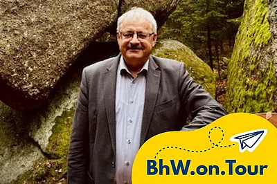 Ein Mann steht lächelnd vor einer großen, moosbedeckten Felsformation in einem Wald. Er lehnt an einer Holzbank mit einem herzförmigen Schild, das einen freundlichen Akzent in der natürlichen Umgebung setzt.