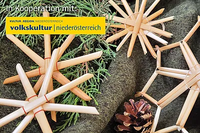 Strohsterne liegend auf einem grauen Hintergrund. Daneben liegen ein paar grüne Zweige und ein kleiner Tannenzapfen.