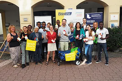 Gruppenbild auf der Straße vor dem BhW. Man sieht eine Gruppe von 16 Personen. Dabei sind Trainer/innen der Basisbildung, Schauspieler Sven Kaschte und Passanten. Sie halten kleine Sackerl mit Buchstaben-Nudeln und einem Info-Zettel über den Weltalphabetisierungstag in den Händen.