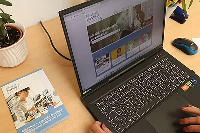 Auf einem Schreibtisch steht ein aufgeklappter Laptop und daneben liegt eine Broschüre von der Bildungsdatenbank. Auf der Tastatur des Laptops tippen zwei Hände. Auf dem Bildschirm ist die Bildungdatenbank zu sehen.
