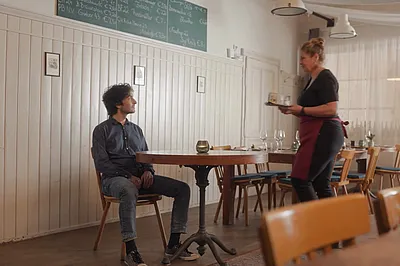 Ein Mann sitzt im Wirtshaus an einem Tisch. Eine Kellnerin steht mit einem Tablett und einer Tasse vor ihm.