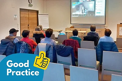 Mehrere Personen sitzen in einem Seminarraum und verfolgen eine Online-Präsentation, die auf eine Leinwand projiziert wird. Auf dem Bildschirm ist eine Frau im Videocall mit dem Logo von „BhW Niederösterreich“ zu sehen.