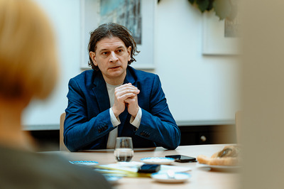 Gehörlosenverband Niederösterreich-Leiter Lukas Huber sitzt an einem Tisch mit Glas, Kaffeegeschirr und Kuchenteller darauf und verschränkt die Hände. Ihm gegenüber und mit dem Rücken zur Kamera sitzt eine Frau mit blondem Haar.