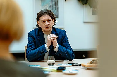Gehörlosenverband Niederösterreich-Leiter Lukas Huber sitzt an einem Tisch mit Glas, Kaffeegeschirr und Kuchenteller darauf und verschränkt die Hände. Ihm gegenüber und mit dem Rücken zur Kamera sitzt eine Frau mit blondem Haar.