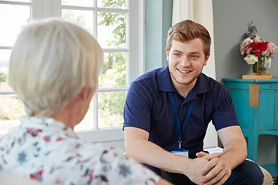 Ältere Frau im Gespräch mit einem jungen Pfleger auf einem Sofa, er lächelt sie an.