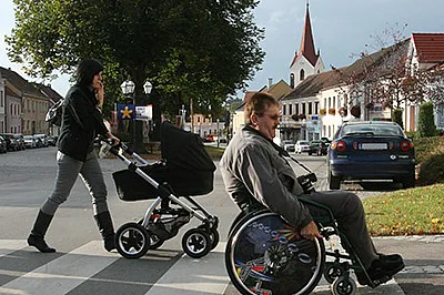 eine Frau mit Kinderwagen und ein Rollstuhlfahrer überqueren eine Straße auf einen Zebrastreifen