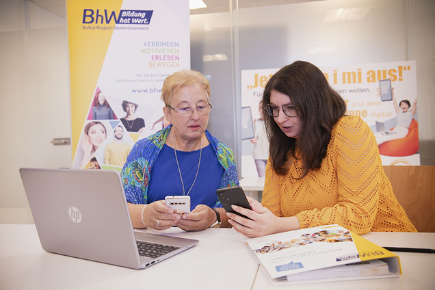 Zwei Frauen sitzen gemeinsam an einem Tisch, schauen konzentriert auf ein Smartphone und tauschen sich über dessen Nutzung aus. Vor ihnen liegen ein Laptop und Unterlagen. Im Hintergrund sind bunte Roll-Ups mit Bildungsbotschaften und fröhlichen Bildern zu sehen.