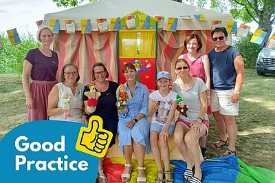 Eine Gruppe von acht Personen sitzt und steht vor einem bunten Zelt, das mit BhW-Wimpeln geschmückt ist, und einige halten Handpuppen in den Händen. Im linken unteren Bildbereich befindet sich ein blaues „Good Practice“-Grafikelement mit einem Daumen-hoch-Symbol.