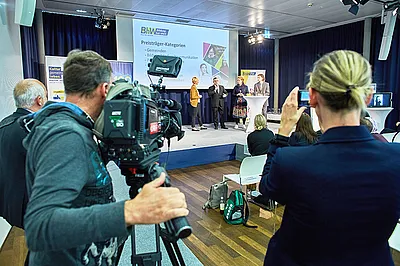 Im Vordergrund sieht man einen Mann mit einer großen Fernsehkamera und daneben eine Frau mit einem Handy mit dem sie filmt. beide filmen eine Interviewszene auf einer Bühne, auf der vier Personen stehen.