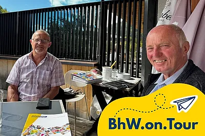 Zwei Männer sitzen lächelnd an einem Tisch in einem sonnigen Außenbereich eines Cafés und haben BhW-Unterlagen vor sich liegen. Im Vordergrund ist ein gelbes BhW.on.Tour-Grafikelement mit Papierflieger-Symbol eingeblendet.