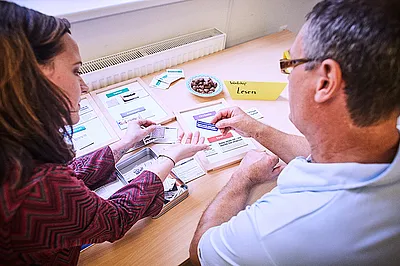 Zwei Personen sitzen an einem Tisch und sortieren gemeinsam Karten mit Texten. Die Szene zeigt eine Lernsituation, in der Inhalte besprochen und zugeordnet werden, unterstützt durch übersichtliche Materialien und eine ruhige Arbeitsatmosphäre.