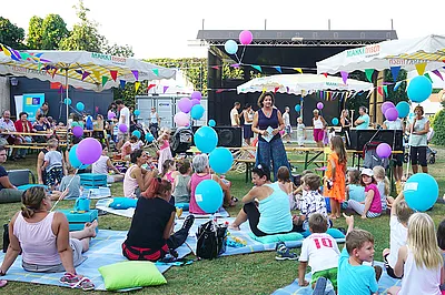 Auf dem Bild ist das Setting eines Lesepicknicks im Freien zu sehen, mit Picknickdecken, Kindern und ihren Eltern sowie eine Bühne für eine Lesung.