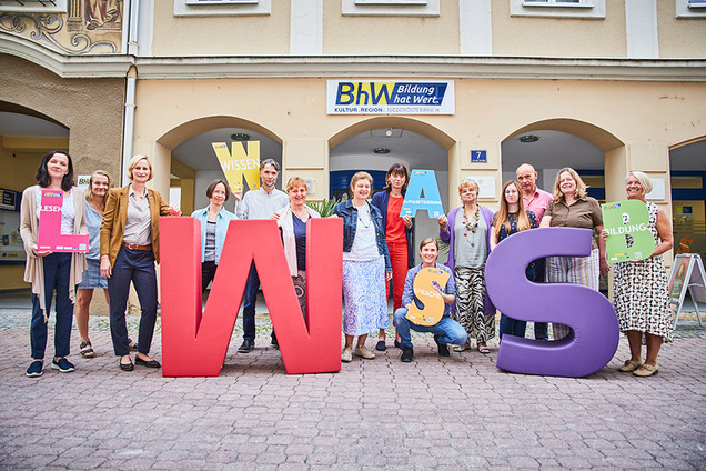 Die Lernbegleiterinnen und Lernbegleiter der Basisbildung stehen vor dem Haus Linzerstraße 7. Davor stehen noch zwei sehr große Buchstaben aus Schaumstoff. Ein großes "W" in rot und ein großes "S" in lila. Einige Personen halten auch kleinere Buchstaben in den Händen.