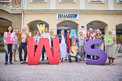 Die Lernbegleiterinnen und Lernbegleiter der Basisbildung stehen vor dem Haus Linzerstraße 7. Davor stehen noch zwei sehr große Buchstaben aus Schaumstoff. Ein großes "W" in rot und ein großes "S" in lila. Einige Personen halten auch kleinere Buchstaben in den Händen.