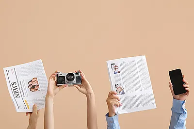 Das Bild zeigt einige hochgestreckte Arme, die Zeitungen, eine Kamera und ein Smartphone halten.