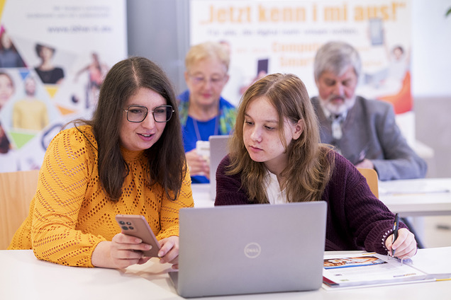 In einem Workshop sitzen mehrere Personen an Tischen und arbeiten gemeinsam an Laptops und Smartphones. Im Vordergrund unterstützt eine junge Frau eine Schülerin bei der digitalen Anwendung. Im Hintergrund beschäftigen sich weitere Teilnehmende konzentriert mit ihren Geräten, um ihre digitale Kompetenzen zu stärken.