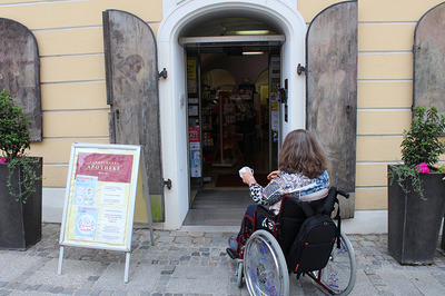 Eine Frau im Rollstuhl vor dem stufenlosen Eingang zu einer Apotheke