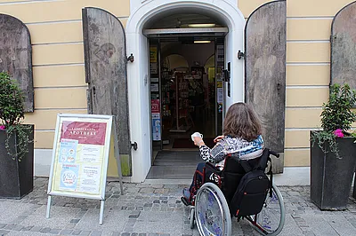 Eine Frau im Rollstuhl vor dem stufenlosen Eingang zu einer Apotheke