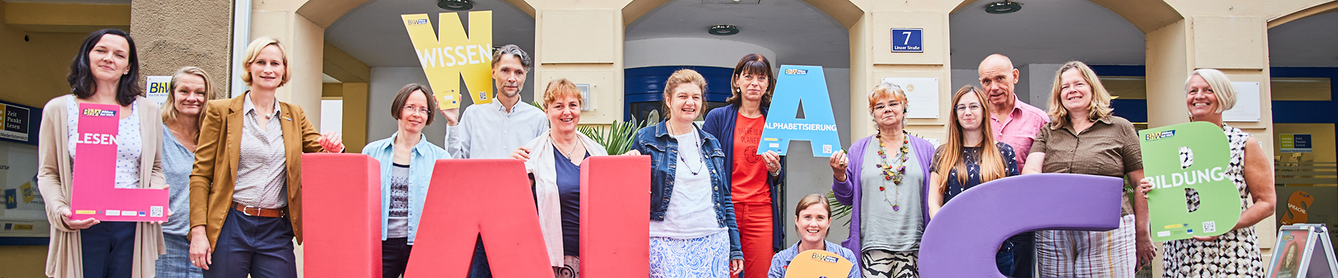 Die Lernbegleiterinnen und Lernbegleiter der Basisbildung stehen vor dem Haus Linzerstraße 7. Davor stehen noch zwei sehr große Buchstaben aus Schaumstoff. Ein großes "W" in rot und ein großes "S" in lila. Einige Personen halten auch kleinere Buchstaben in den Händen.