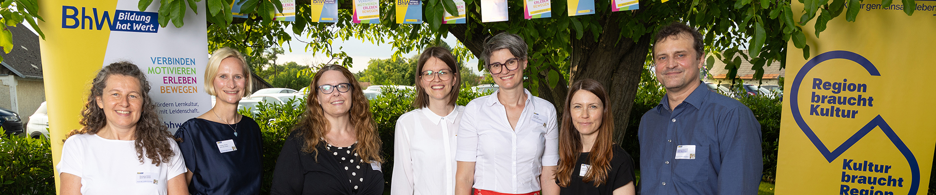 Auf dem Foto stehen sieben Personen lächelnd nebeneinander im Freien, zwischen zwei Roll-Ups mit den Logos von BhW Bildung hat Wert und Kulturregion Niederösterreich. Im Hintergrund hängt eine bunte Wimpelkette, und die Gruppe wirkt freundlich und professionell.